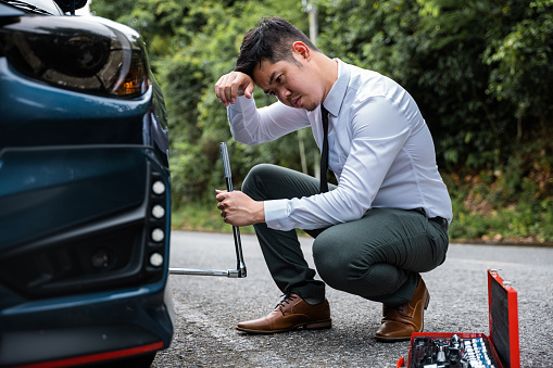 Confident young man in uniform changing car tyre on the roadside. Roadside assistance by Asian businessman wearing white shirt. Expert car mechanic at work.