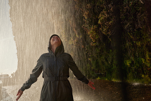 Young carefree woman in raincoat having fun while standing under the waterfall in nature.