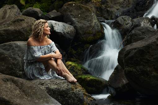 Carefree woman day dreaming while sitting on the rock by the waterfall in nature.