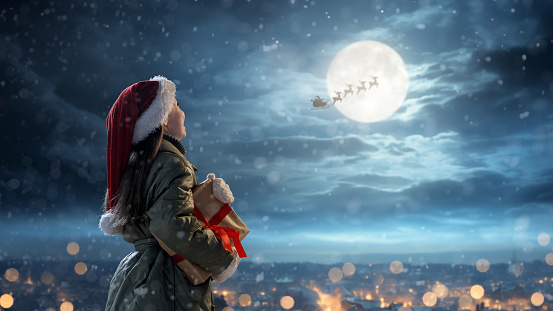 Merry Christmas. Cute little child with xmas present. Santa Claus flying in his sleigh against moon sky. Happy kid enjoy the holiday. Portrait of girl with gifts on dark background.