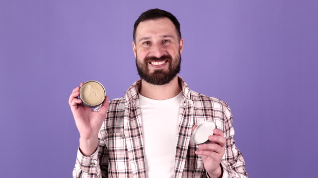 Handsome bearded man holding hair wax, clay on purple background.