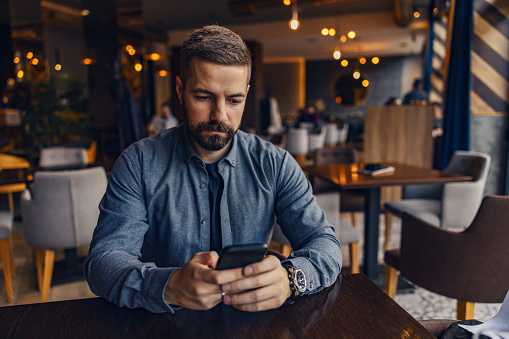 A serious young man is sitting in cafe and ordering drinks on mobile app.