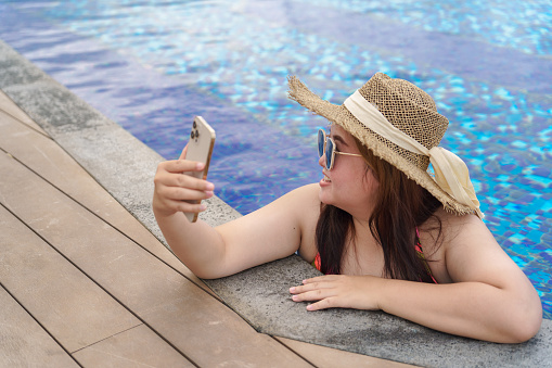 Overweight young woman orange swimsuitÂ relaxing and holding smartphone in swimming pool. Obese Woman Vacation Traveling hand using phone.
