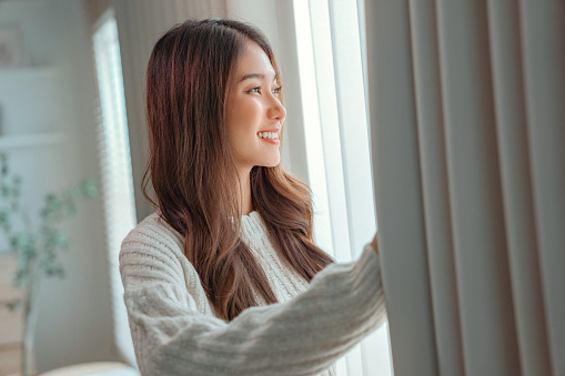 Close up of happy young asian woman opening curtain at home, Smiling attractive lady looking outside apartment enjoying dreaming and contemplating feeling hope concept