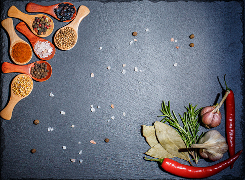 Spices in wooden spoons and cherry tomatoes, chili peppers, garlic and herbs for cooking on a black surface and black background