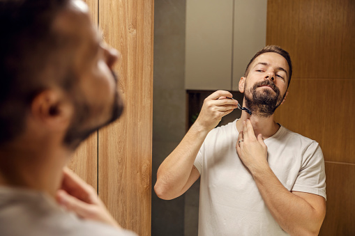 Reflection of a young bearded man shaving his beard with razor blade in a bathroom.
