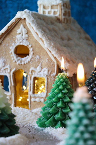 Stock photo showing close-up view of a group of moulded Christmas tree design wax candles displayed with a gingerbread house in a snowy, Christmas scene.