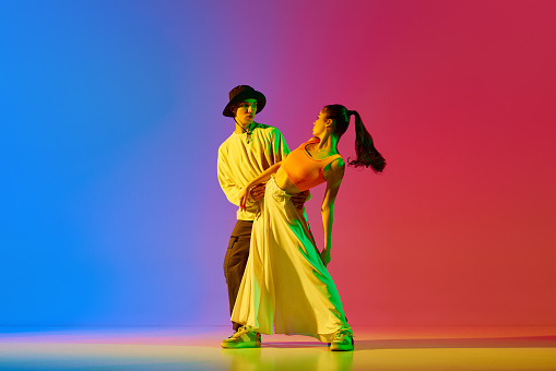 Stylish, young, active and talented boy and girl in modern clothes dancing hip-hop against blue red background in neon light. Concept of hobby, action, street style, contemporary dance, youth, fashion