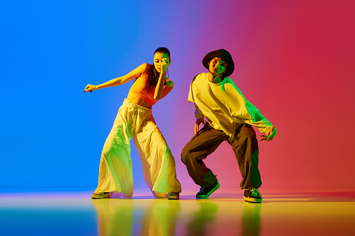 Expressive, talented, artistic young man and woman, hip hop dancers in motion, performing over blue red background in neon. Concept of hobby, action, street style, contemporary dance, youth, fashion