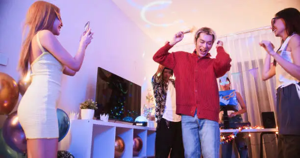 Photo of Group of young diverse millennial Asian friends with LGBTQIA+ people dance together at home party, celebrate New Year, Christmas, or Birthday at night. Happy celebration event, fun activity concept