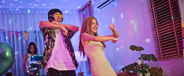 Young millennial Asian duo friends, man and woman dance together at home party, celebrate New Year, Christmas, or Birthday at night. Happy people celebration event, fun activity concept