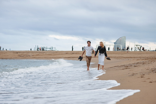 A Caucasian woman and a Spanish man are walking barefoot and holding their shoes at the beach in Barcelona. At the back there are some buildings and tourists also enjoying the day.