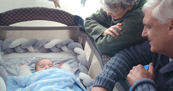 Love, watching and grandparents with sleeping baby in cot in modern family home together babysitting. Happy, sweet and senior people in retirement bonding with boy child, newborn or kid at house.