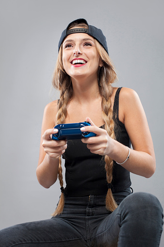 Cheerful young woman in hat and pigtail hairstyle playing console with joystick on gray background looking away. Video game.