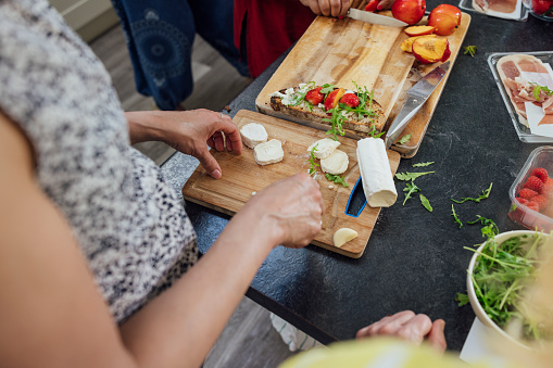 An overhead view of a woman cutting up cheese on a cutting board. A sandwich and other food products lie around the table.