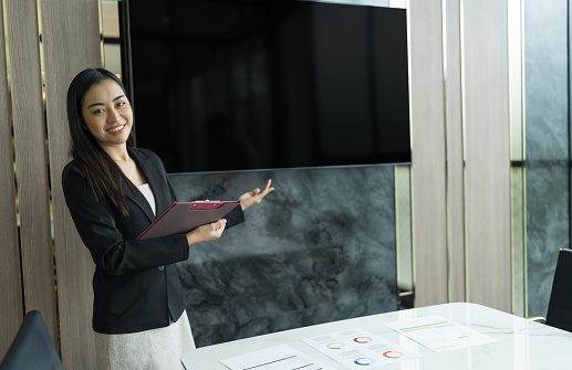 BeautifulÂ Asian Business Woman in black suit pointingÂ to present screen Wall TV ShowingÂ Digital Entrepreneur presenting a project on emptyÂ blank screen TV mock up presentation