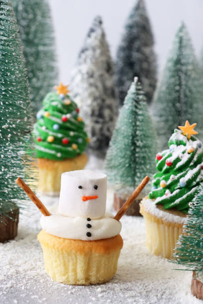 Close-up image of individual, homemade, snowman design cupcake displayed in conifer forest Christmas scene, Christmas tree design cupcakes, plastic model fir trees, icing sugar snow, pale blue background, focus on foreground