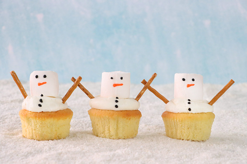 Stock photo showing close-up view of a row of three, freshly baked, homemade snowman design cupcakes, displayed on icing sugar snow against a pale blue background. The cakes are topped with swirls of white butter icing and decorated with large white marshmallows and salted pretzel stick arms. Home baking concept.