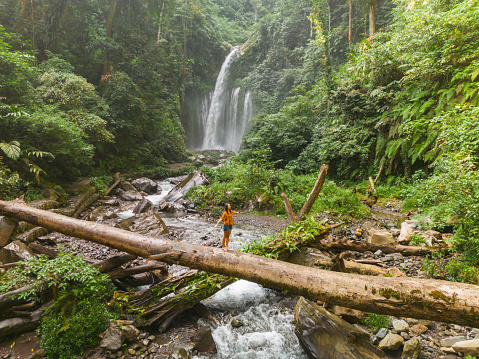 Aerial view of woman crossing river by log on the background of tropical waterfall while hiking in jungles
