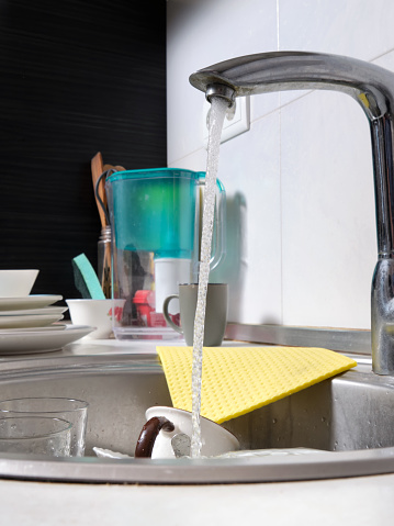 Close-up of a kitchen sink with dirty dishes. Water flows from an open tap. Reluctance to do routine housework. Vertical image.