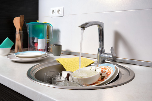 Close-up of a kitchen sink with dirty dishes. Water flows from an open tap. Reluctance to do routine housework after dinner.