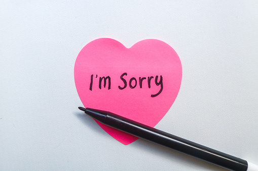 The word sorry written on a pink sticky note in the shape of a heart isolated on a white background. Concept of Apologizing with note and pen