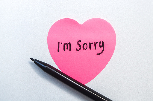 The word sorry written on a pink sticky note in the shape of a heart isolated on a white background. Concept of Apologizing with note and pen