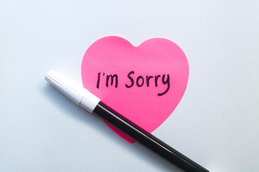 The word sorry written on a pink sticky note in the shape of a heart isolated on a white background. Concept of Apologizing with note and pen