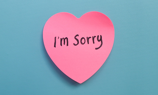 The word sorry written on a pink sticky note in the shape of a heart isolated on a blue background. Concept of Apologizing with note