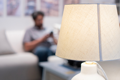 Close up of lamp spending warm light at cozy reader's corner with blurred of man sit on sofa behind. Selective focus, copy space.