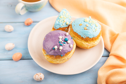Purple and blue glazed donut and cup of coffee on blue wooden background and orange linen textile. side view, selective focus. Breakfast, morning, concept.