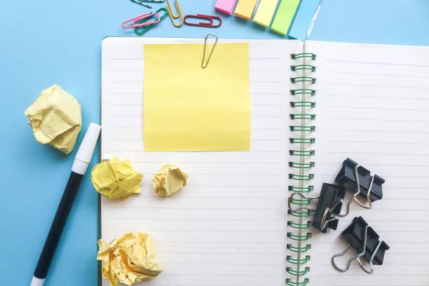 istock Yellow blank sticky notes on top of empty notebook and isolated on blue background with other objects like pen, paper clip, binder clip, crumpled paper. Open notepad with office supplies 1803894487