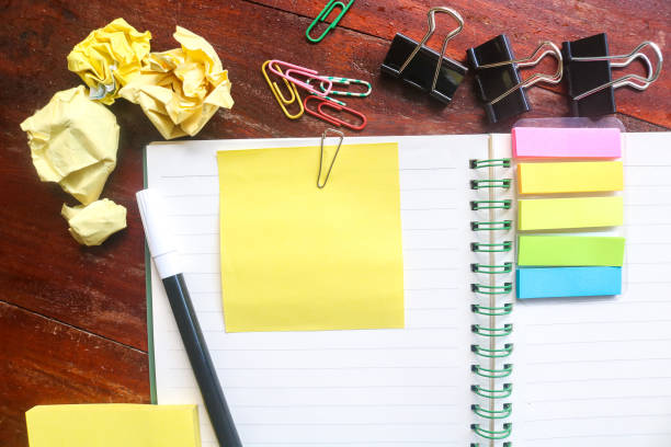 istock Blank yellow sticky notes on top of an empty notebook and isolated on a messy wooden table with other objects like pens, paper clips, binder clips, crumpled paper. Open notepad with office supplies 1803895232