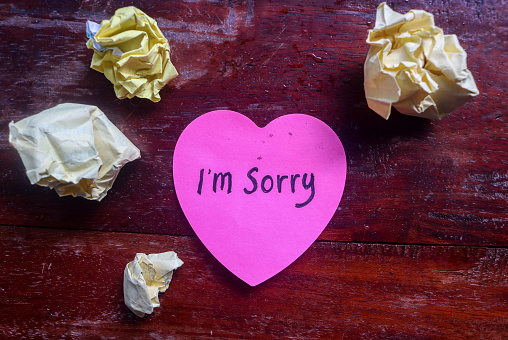 The word sorry written on a pink sticky note in the shape of a heart isolated on a table background and crumpled paper. Concept of Apologizing with note