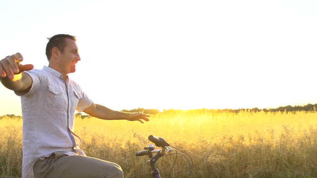 HD1080p: TWO SUPER SLOW MOTION shots of a cheerful man cycling without hands along wheat field on a sunny day.