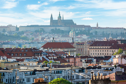 Panoramic photo of the  Prague Castle and the surrounding city. The castle is a large Gothic structure with multiple spires and towers. It is located on a hill overlooking the city. The city is made up of a mix of old and new buildings, with many red roofs. The sky is blue with white clouds. The photo is taken from a high vantage point, looking down on the city.