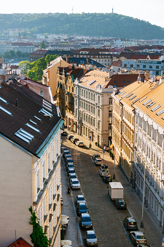 A narrow and winding cobblestone street in Prague, Czech Republic. The street is lined with parked cars and historic buildings with red roofs. The buildings are mostly beige and white in color. In the background, you can see the city’s skyline and hills. The photo was taken during the day, with sunlight shining on the buildings.