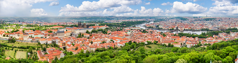 A breathtaking panoramic view of Prague from Petrin Hill, including the Prague castle. The city’s red rooftops and the Vltava River are visible in the distance, while the greenery of the hill is in the foreground. The sky is a beautiful shade of blue with white clouds, and the colors are vibrant.