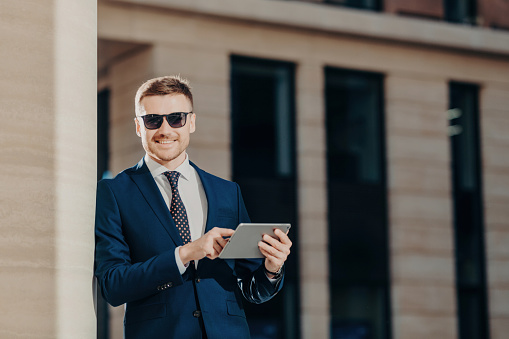 Stylish businessman in sunglasses using a tablet outside modern office buildings on a sunny day