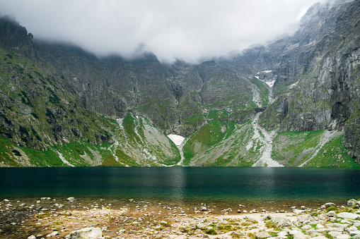 Scenic view of foggy mountains cover by dark clouds and green forest with a reflection in a lake. Stony shore. Morskie Oko. High Tatras, Zakopane, Poland