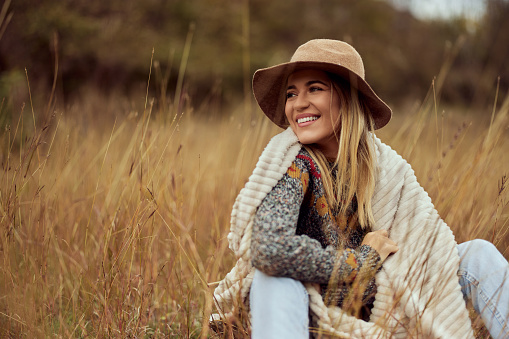 A beautiful blonde girl, sitting in the field, looking away, wearing a hat, dressed nice.