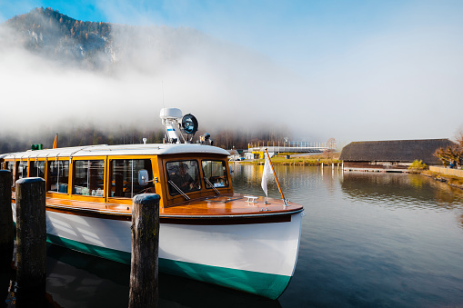 Small boat moored near wooden pier on lake Konigsee in Berchtesgaden. Morning winter fog over mountains on background.