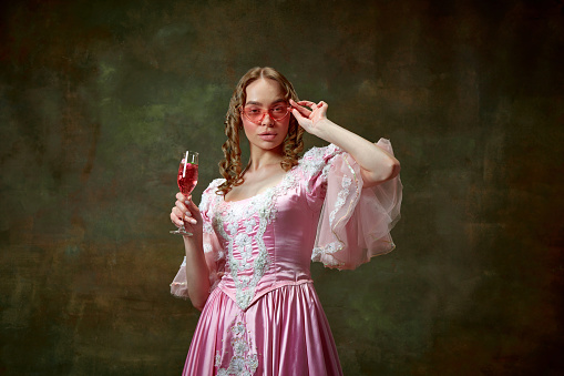 Stylish princess. Portrait of beautiful blond princess wearing pink dress and sunglasses holding wine glass over vintage texture background. Concept of medieval, beauty, old-fashioned clothes, ad