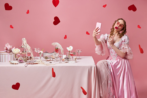 Valentines day, card, red hearts. Portrait of beautiful blond princess, queen wearing fancy pink dress and taking a selfie on cell phone. Concept of love, relationship, medieval, beauty, romance
