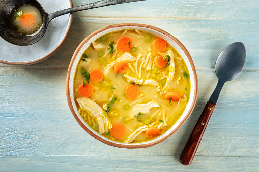 Chicken noodle soup with vegetables, a bowl of healthy stock with a ladle, top shot on a rustic wooden table