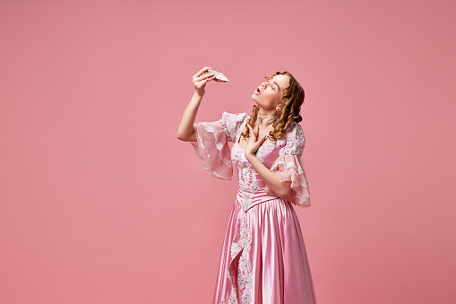 Queen with seafood. Shot of beautiful young princess wearing pretty dress and eating oysters with pleasure over pink studio background. Concept of food, beauty, medieval, fashion, banquet, ad