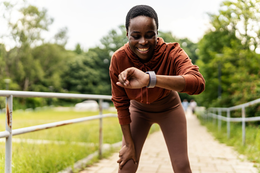 One beautiful african american female athlete checking her smartwatch while exercising outdoors. A young athletic woman smiling while tracking her progress on a fitness watch during her workout.