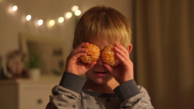 Merry Christmas. Funny child boy laughing and dancing covering his eyes with tangerine. Happy New Year. Portrait of excited little kid wearing pajamas having fun at home on Xmas night. Festive mood