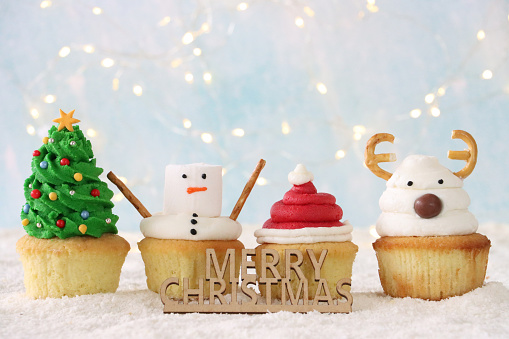 Stock photo showing close-up view of a Merry Christmas sign in front of a row of four, freshly baked, homemade Christmas tree, snowman, Santa hat and reindeer design cupcakes, displayed surrounded by illuminated fairy lights on icing sugar snow, against a pale blue background. Home baking concept.