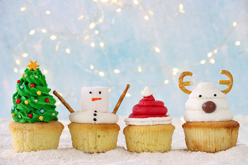 Stock photo showing close-up view of a row of four, freshly baked, homemade Christmas tree, snowman, Santa hat and reindeer design cupcakes, displayed surrounded by illuminated fairy lights on icing sugar snow, against a pale blue background. Home baking concept.